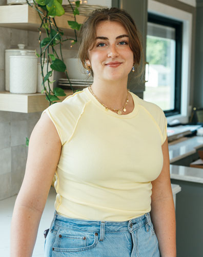 Young woman with brown hair smiling. She is wearing a yellow shirt and jeans.